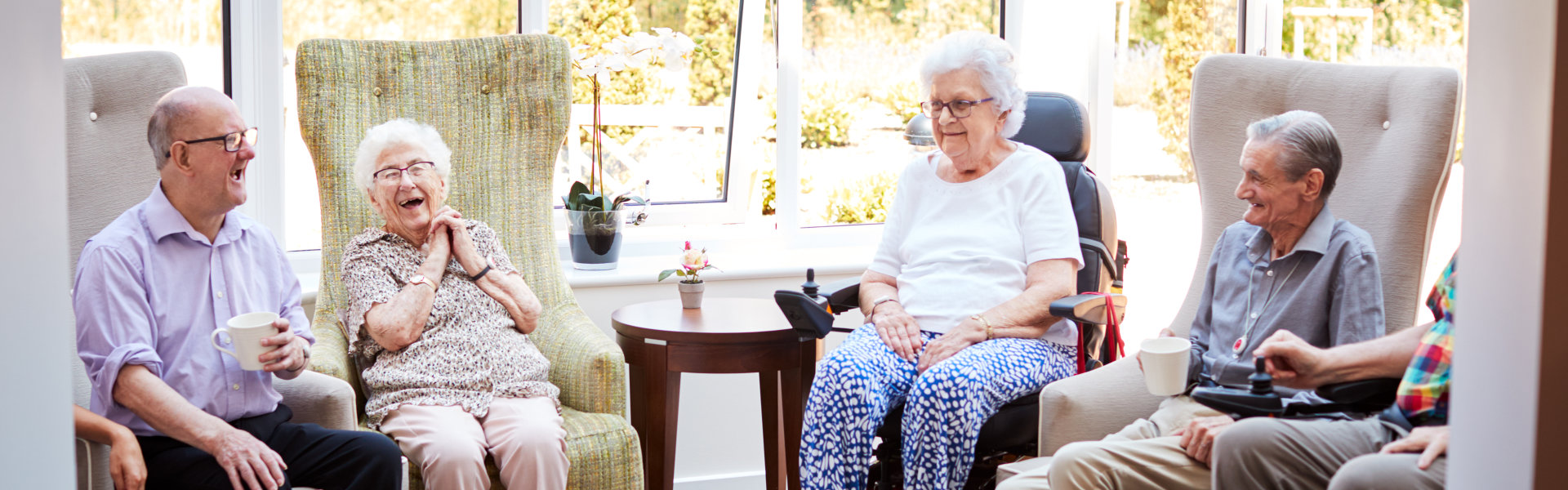 Male And Female Residents Sitting In Chairs And Talking With Carer In Lounge Of Retirement Home